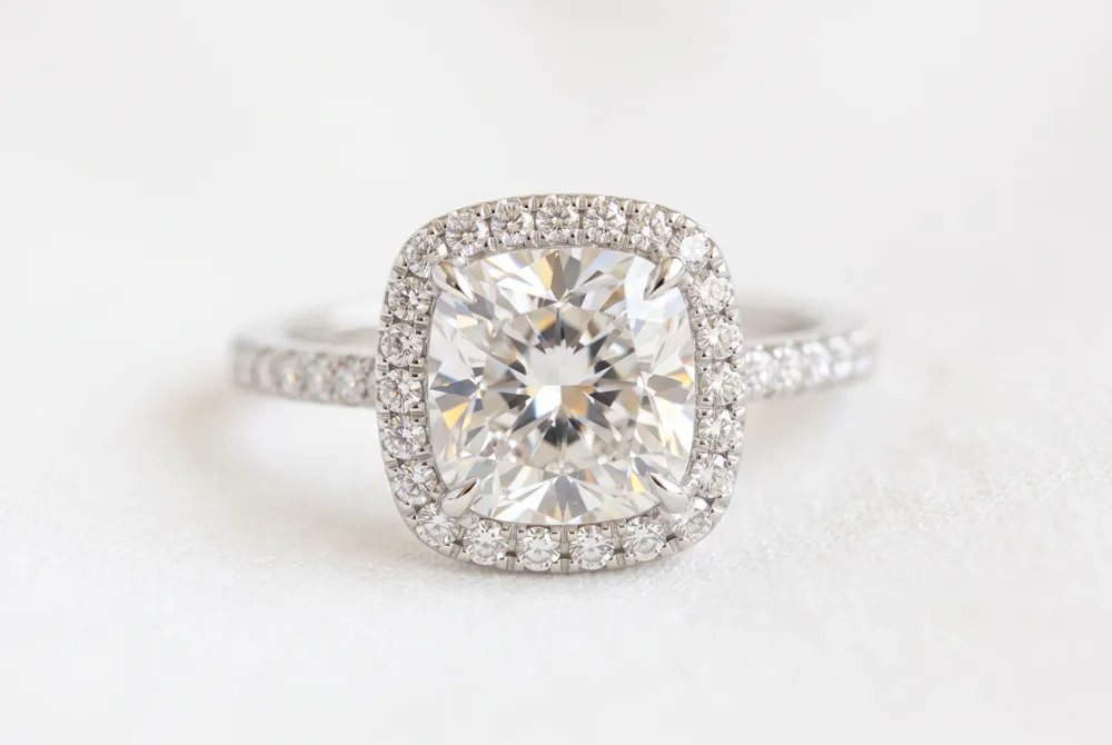 A halo of delicate moissanite stones encircles the cushion cut, amplifying its brilliance and creating an illusion of a larger, more commanding presence that catches the light from every angle.