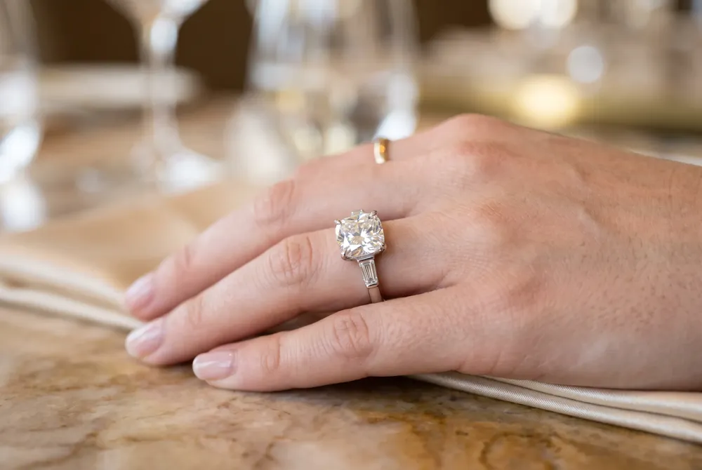 Tapered baguettes flanking a cushion cut center stone create a balanced, symbolic design that enhances the moissanite's fire, making it a meaningful and radiant choice for an engagement ring.