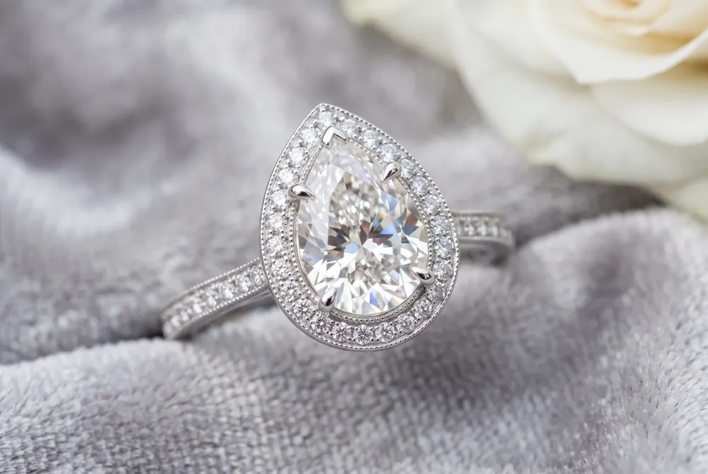 A halo of accent diamonds surrounds the center pear, making it appear larger and more luminous while adding a protective metal buffer around the stone's delicate pointed tip.