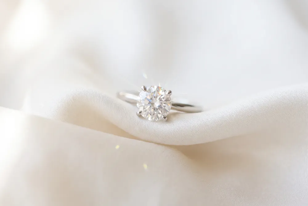 A four-prong solitaire setting allows the cushion cut moissanite's brilliant facets to command attention, its soft corners and radiant sparkle defining understated luxury for the modern bride.